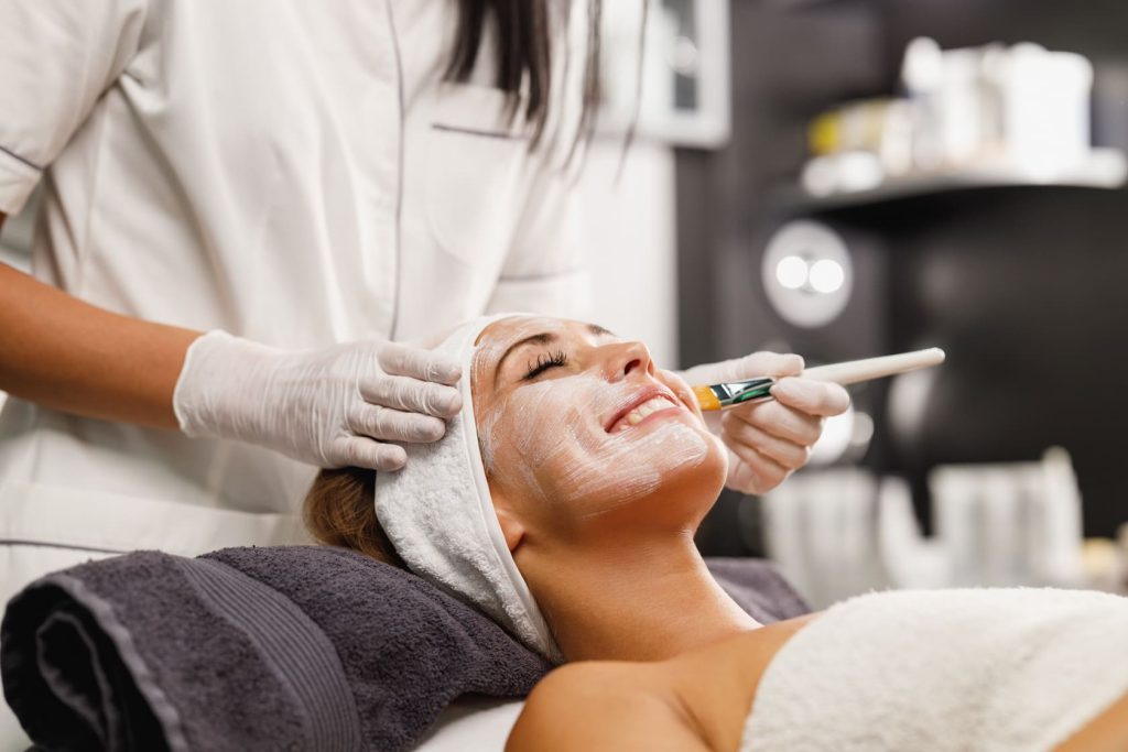 Esteticista aplicando una mascarilla facial a una mujer sonriente recostada en un spa.